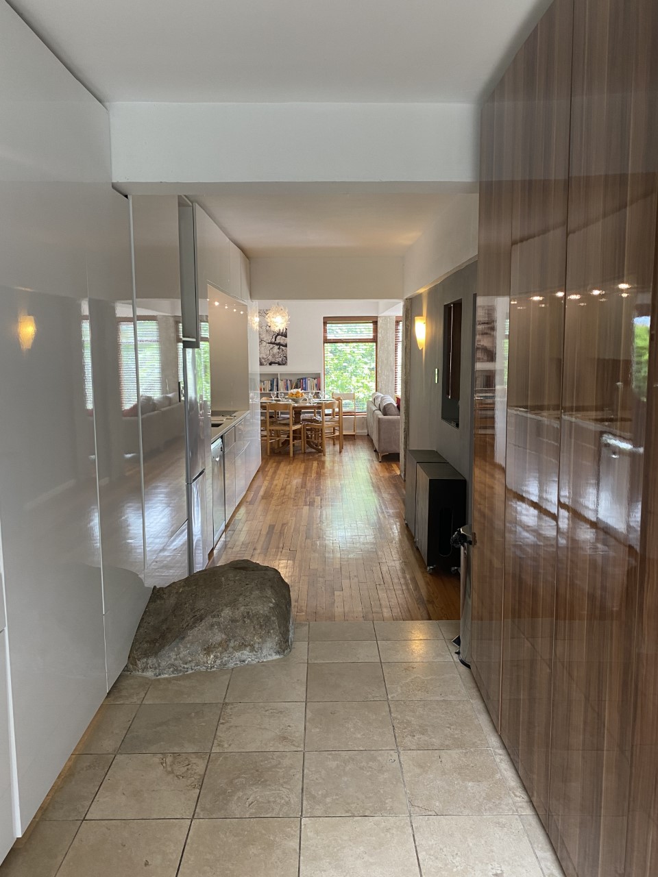 A hallway features a combination of polished wooden and tiled flooring, with a natural stone element present. On one side, sleek cabinetry offers storage, while the view ahead reveals a dining area filled with natural light through large windows.