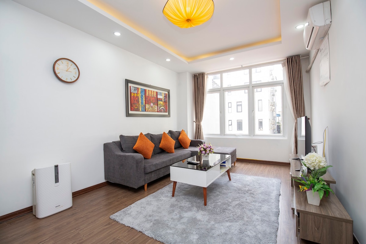 A living area features a gray sofa adorned with orange cushions, positioned across from a low white coffee table. A soft rug covers the wooden floor beneath. Natural light floods through large windows, with a wall clock and framed artwork adding to the ambience.