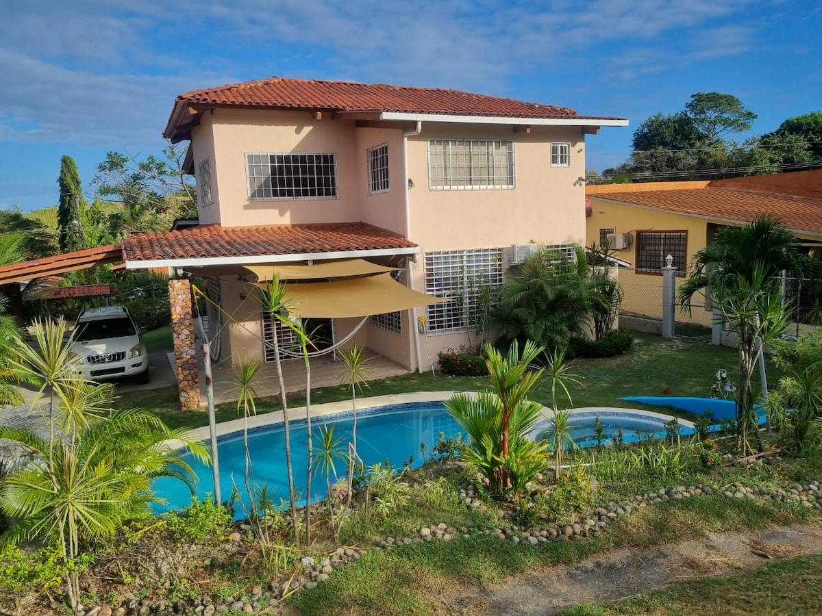 A two-story house with light-colored walls and a terracotta roof is surrounded by lush greenery. A clear blue pool is situated in front, framed by tropical plants. A car is parked under a shaded area, contributing to the relaxed outdoor setting.