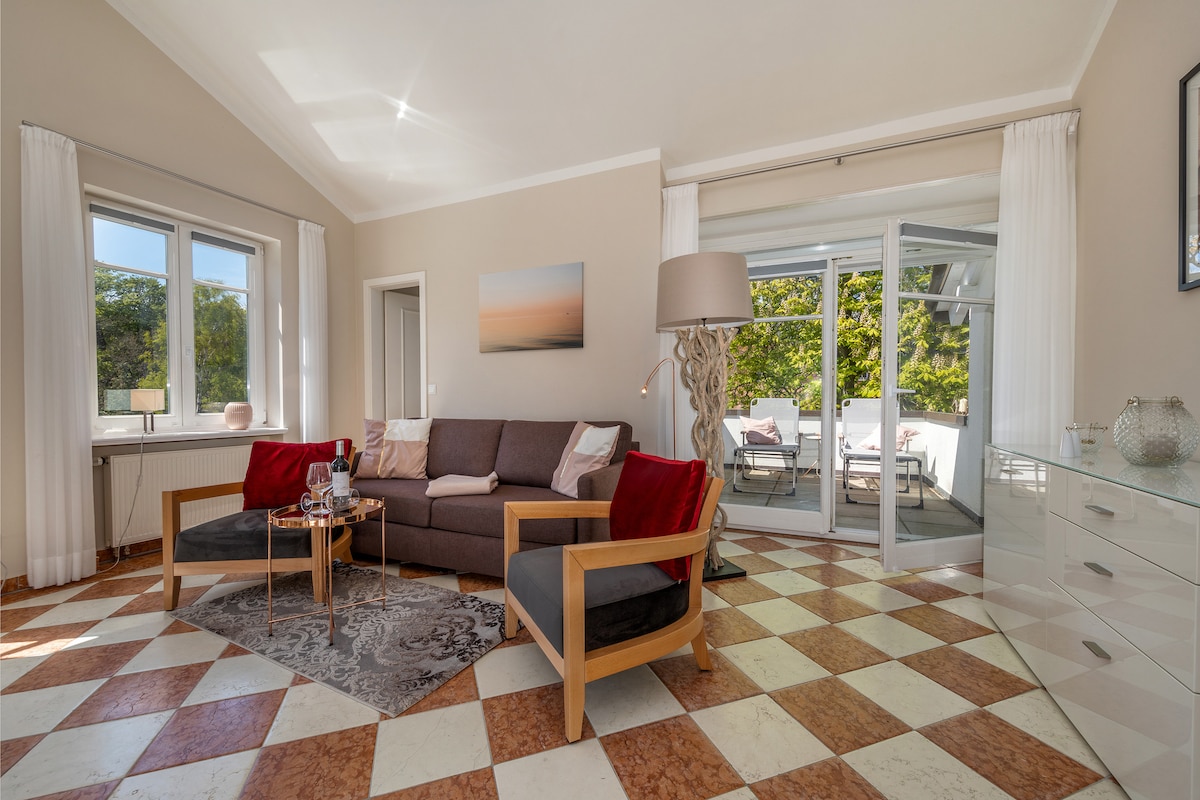 The living area features a cozy seating arrangement with a brown sofa and two wooden armchairs, adorned with red cushions. Large windows offer natural light and provide views of the greenery outside. A decorative lamp stands beside the seating, enhancing the space's inviting feel.