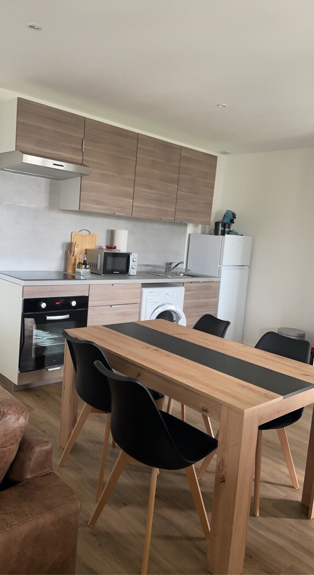 A modern kitchen is displayed, featuring wood cabinetry and a sleek design. A dining table with four chairs is positioned in front. Appliances include an oven, microwave, and refrigerator, alongside a washing machine. Natural light fills the space, creating a functional atmosphere.