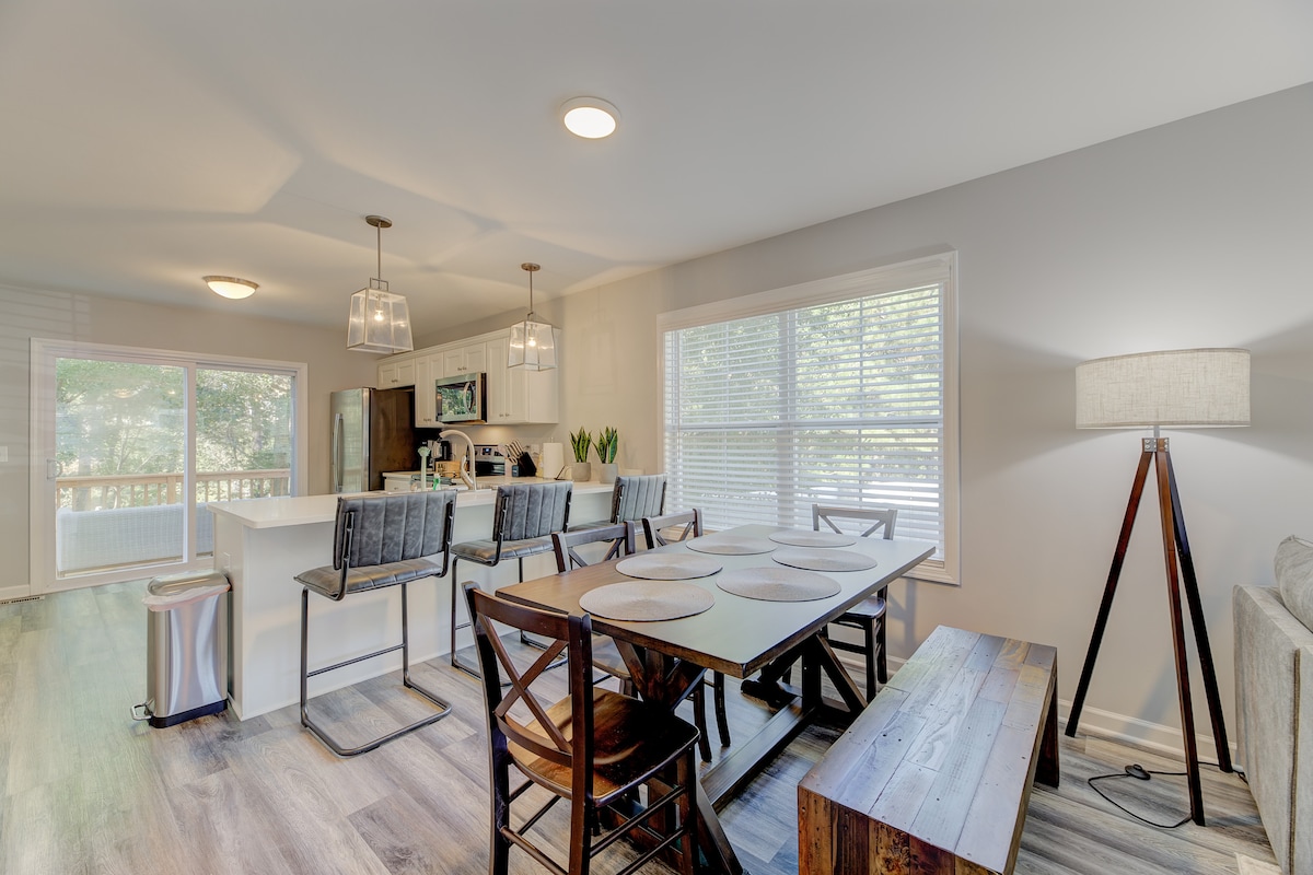 The dining area features a wooden table surrounded by matching chairs, set beside large windows that allow ample natural light. A contemporary lamp stands nearby, illuminating the space, while barstools line the open kitchen counter, creating a welcoming atmosphere.