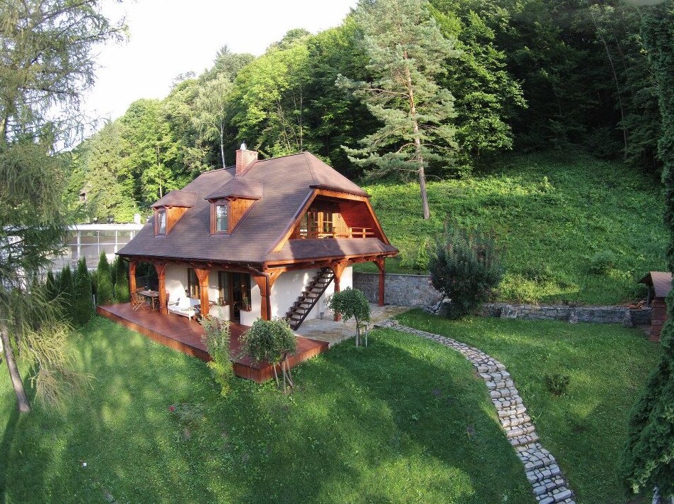 A charming house is set within a lush green landscape, surrounded by trees. The wooden structure features a spacious porch and well-maintained garden path leading to it. A tranquil atmosphere is created by the natural surroundings, inviting guests to relax and enjoy the outdoors.