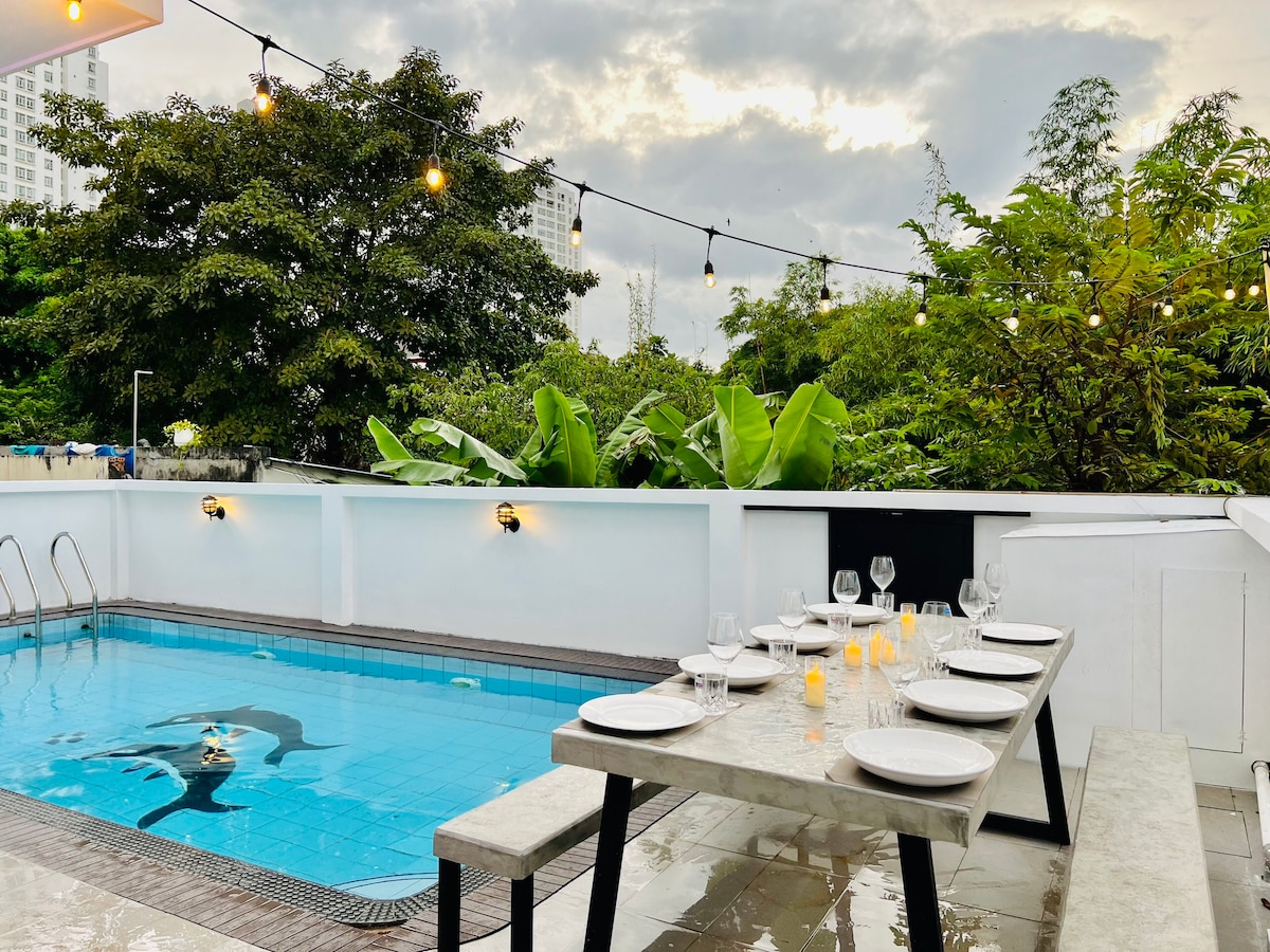 An outdoor dining area is set beside a swimming pool, featuring a long, rectangular table with white plates and glasses arranged on top. Lush greenery surrounds the space, and a string of decorative lights is suspended above, contributing to a serene atmosphere.