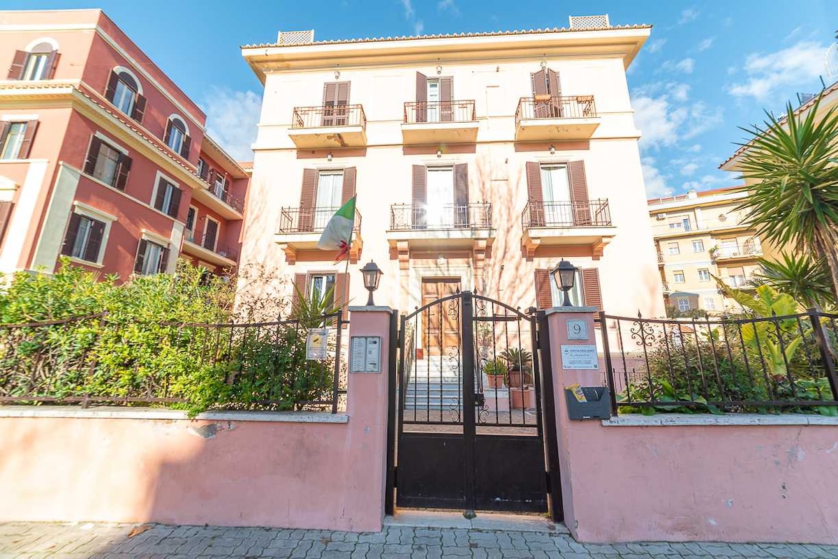A multi-story building is framed by vibrant greenery and a wrought iron gate. The façade features balconies with shutters and decorative elements, while an Italian flag is displayed. A pathway leads to the entrance, inviting guests into the property.