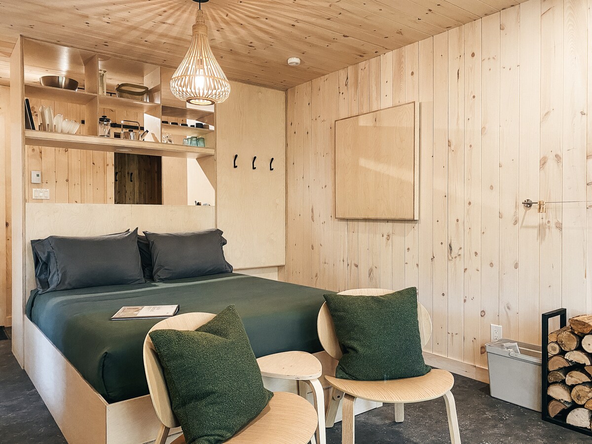 A minimalistic interior features a spacious bed with dark bedding, accompanied by two light wooden chairs and green accent pillows. Warm wood paneling covers the walls, and natural light fills the space, highlighting a cozy atmosphere. A small storage area is visible in the background.