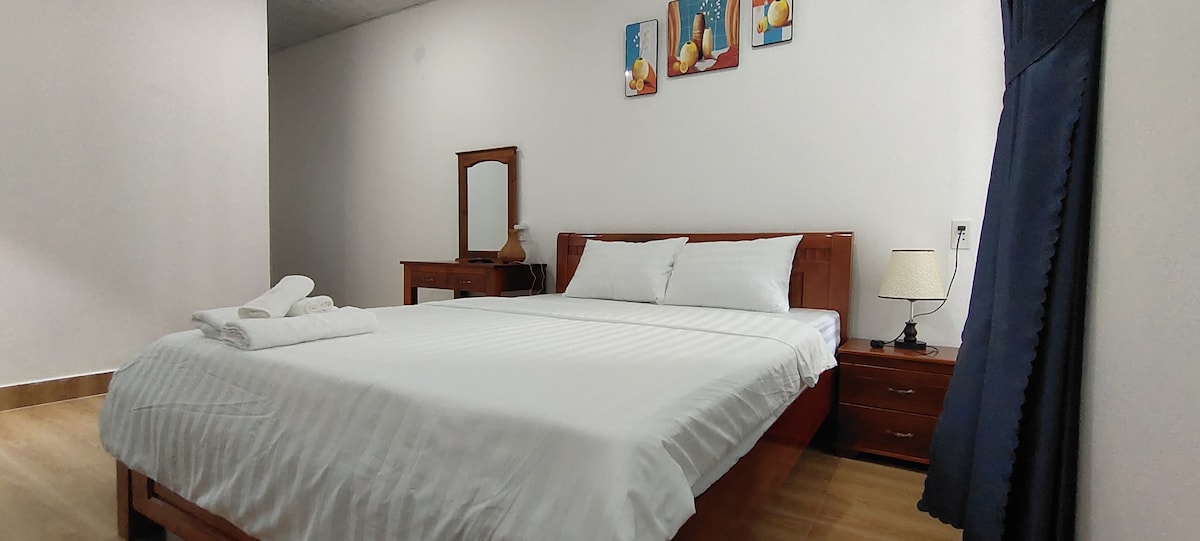 A spacious bedroom is presented with a large bed featuring white linens. A wooden nightstand holds a lamp, while a mirror and a wooden dresser are positioned nearby. Decorative wall art adds a touch of colour to the neutral walls. Light wood flooring completes the room.