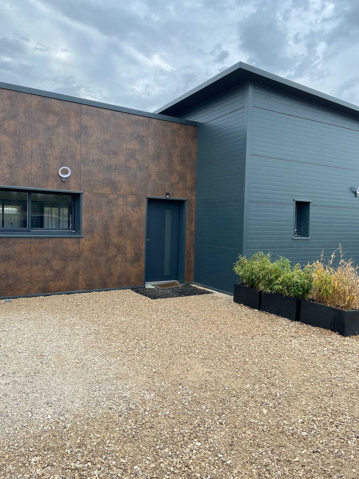 The exterior of the house showcases a modern design with contrasting textures of dark wood and metal. A gravel pathway leads to the entrance, framed by landscaped planters containing greenery. Large windows are visible, enhancing the contemporary appearance.