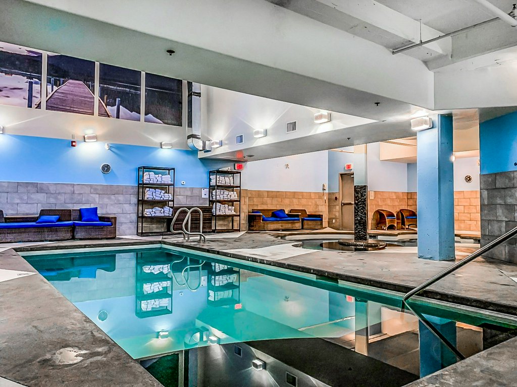 An indoor pool area features a glistening water surface, surrounded by cushioned lounge seating in blue tones. Ambient lighting highlights the textured walls, while a large mural depicting a dock is visible in the background, enhancing the serene atmosphere.