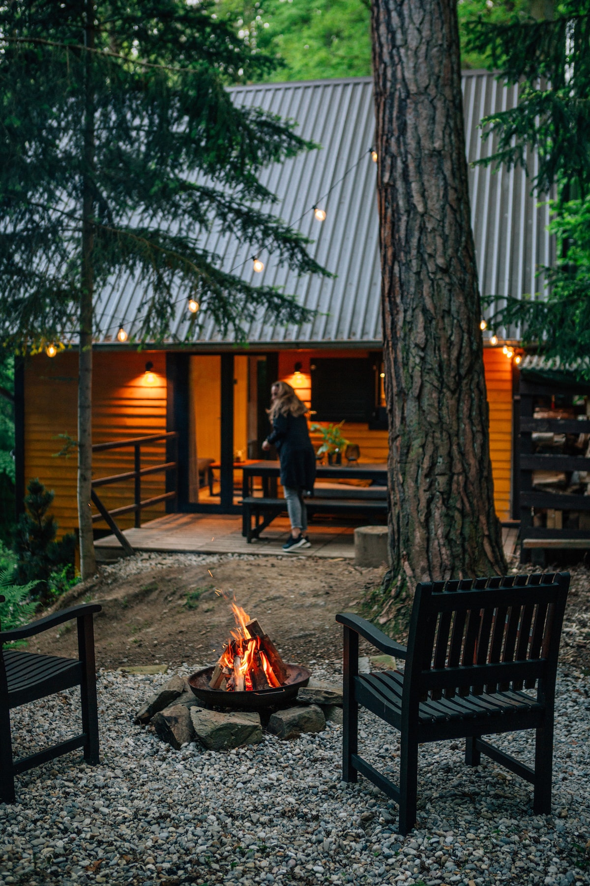 The cabin is nestled among trees, with a cozy outdoor fire pit surrounded by two dark wooden chairs. Soft lighting can be seen from the windows, suggesting a welcoming interior. A gravel path leads to the entrance, enhancing a serene outdoor ambiance.