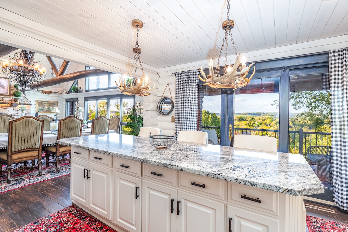 A spacious kitchen features a large granite island with white cabinetry and decorative lighting. Large windows provide an expansive view of the countryside, while rustic elements blend with modern touches. Wooden accents and a vibrant rug enhance the welcoming atmosphere.