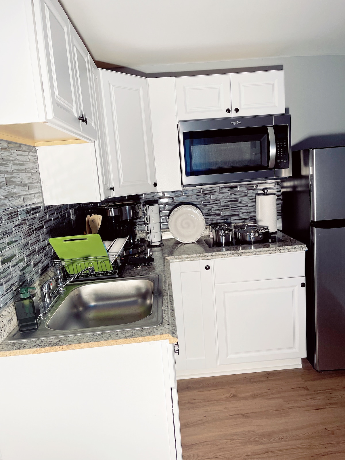 The compact kitchen features white cabinetry and a striking backsplash. A stainless steel microwave is positioned above a gas stove, while a sink with a polished granite countertop completes the area. Various kitchen utensils and dishware are neatly organized, emphasizing functionality.