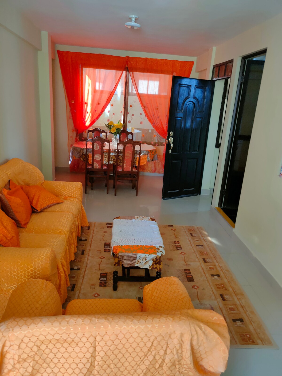 A cozy living area features a set of orange sofas arranged around a low wooden table. A decorative area rug lies beneath, and a dining space with a table and chairs is visible through the open doorway. Bright red curtains add a pop of color.