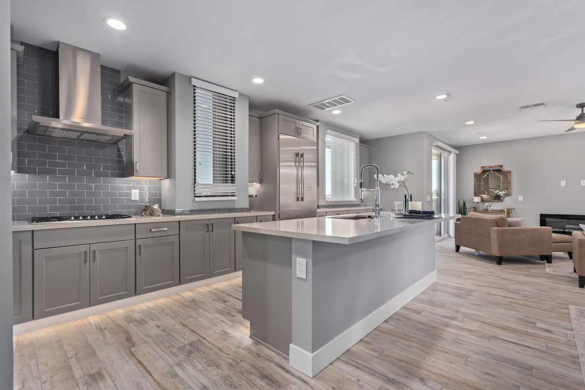 A contemporary kitchen is featured with sleek gray cabinetry and stainless steel appliances. A large island with a smooth countertop provides ample workspace. Natural light streams through the large windows, illuminating the open layout that connects to a comfortable living area.