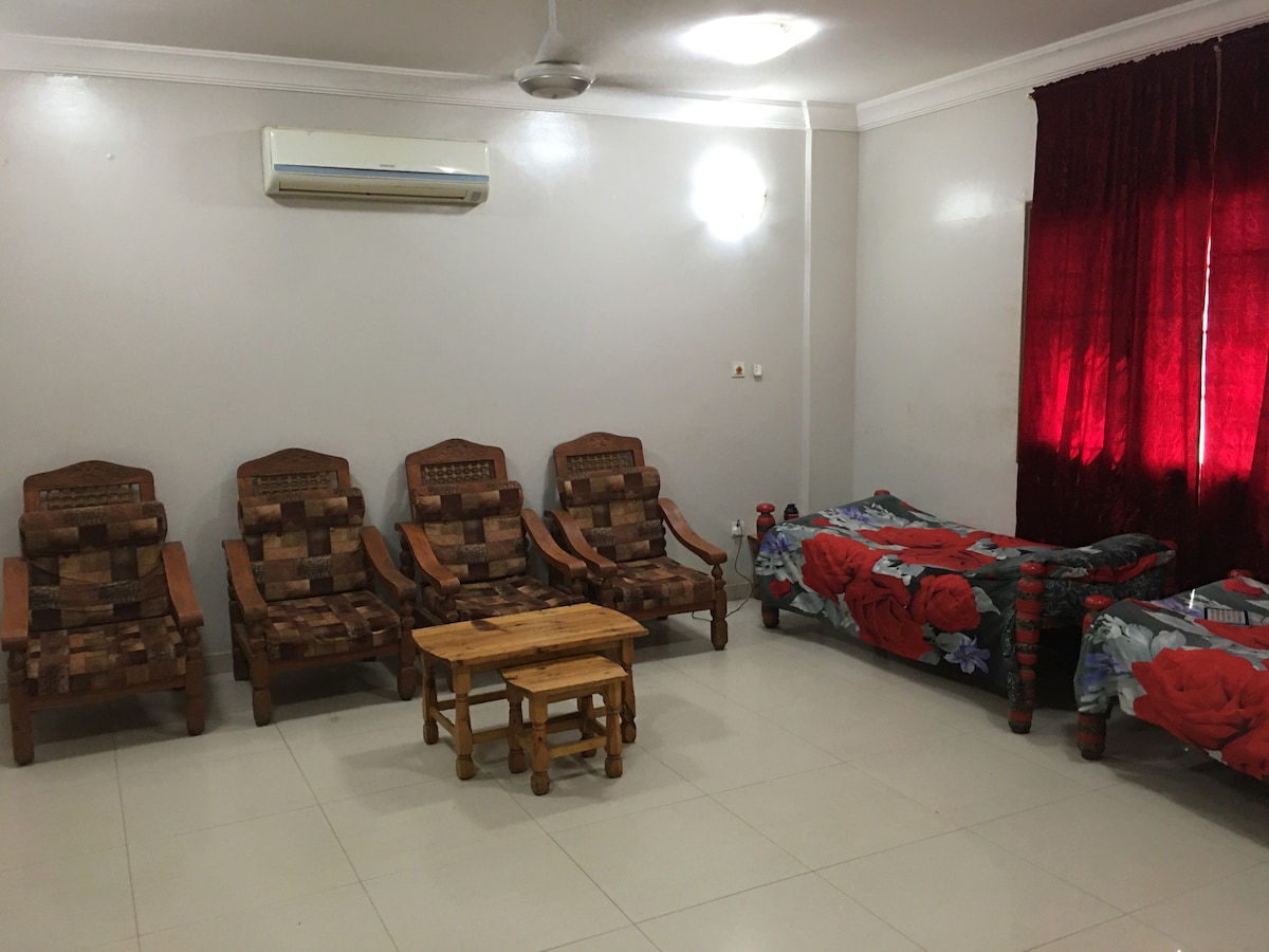 The living area is furnished with four wooden chairs featuring patterned cushions and a small wooden coffee table. A single bed dressed in a floral bedspread is positioned nearby. Natural light enters through a window covered with deep red curtains, complementing the neutral walls.