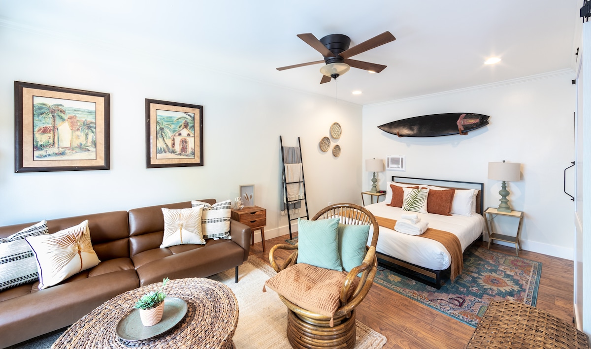 The studio features a brown leather sofa and a round coffee table atop a decorative rug. A queen bed is positioned against the wall, flanked by two lamps. Two framed artworks and a surfboard enhance the decor, while a ceiling fan provides ventilation.