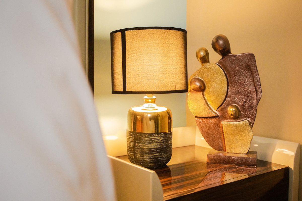 A stylish bedside table is adorned with a sculptural piece and a softly illuminated lamp. The lamp features a round shade that casts a warm glow, while the sculpture, composed of various shapes and materials, adds an artistic touch to the space.