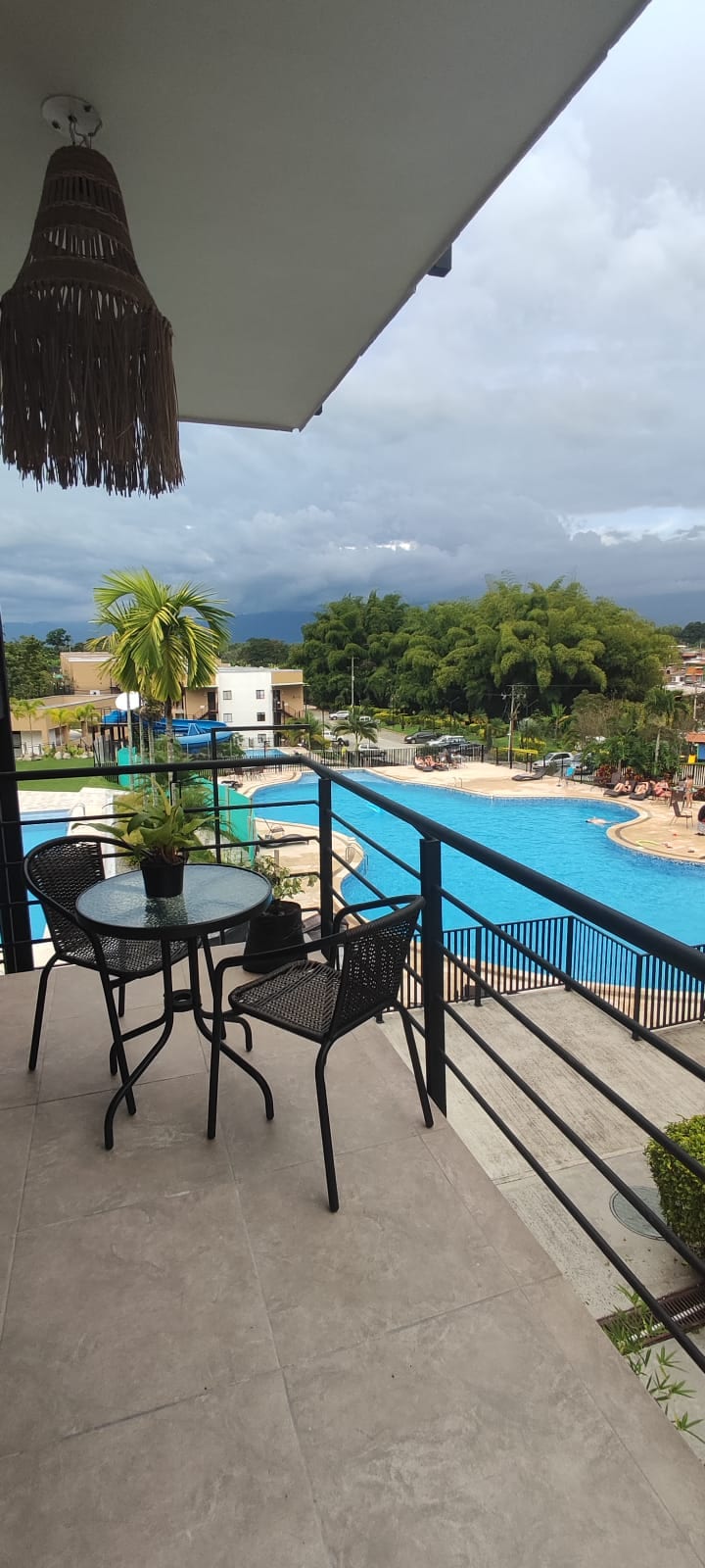 A balcony view showcases a spacious pool area surrounded by greenery and lounge chairs. A small, round table with two chairs is positioned on the balcony, offering a place to relax while enjoying the landscape of the pool and surrounding buildings.