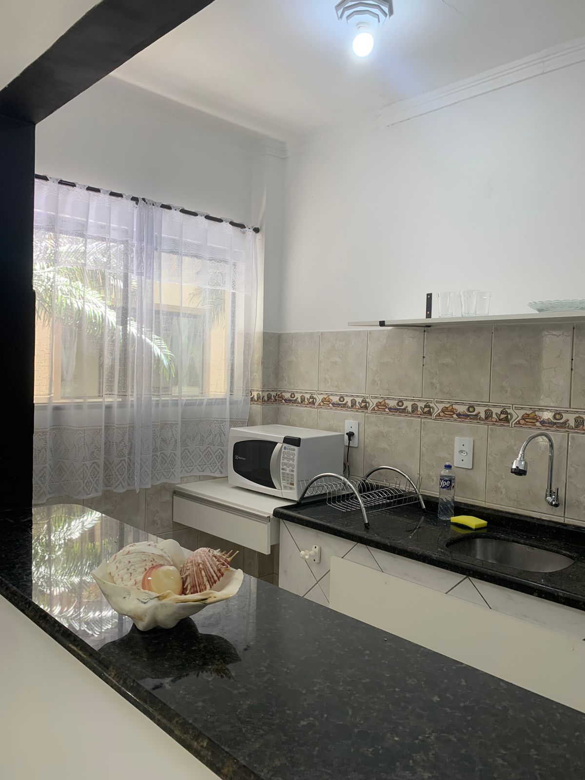 A modern kitchen area is presented, featuring a sleek black countertop and an arrangement of kitchen utensils. Natural light filters through sheer curtains, highlighting a microwave and a sink with a modern faucet. Shelves above the counter display glassware and decorative items.