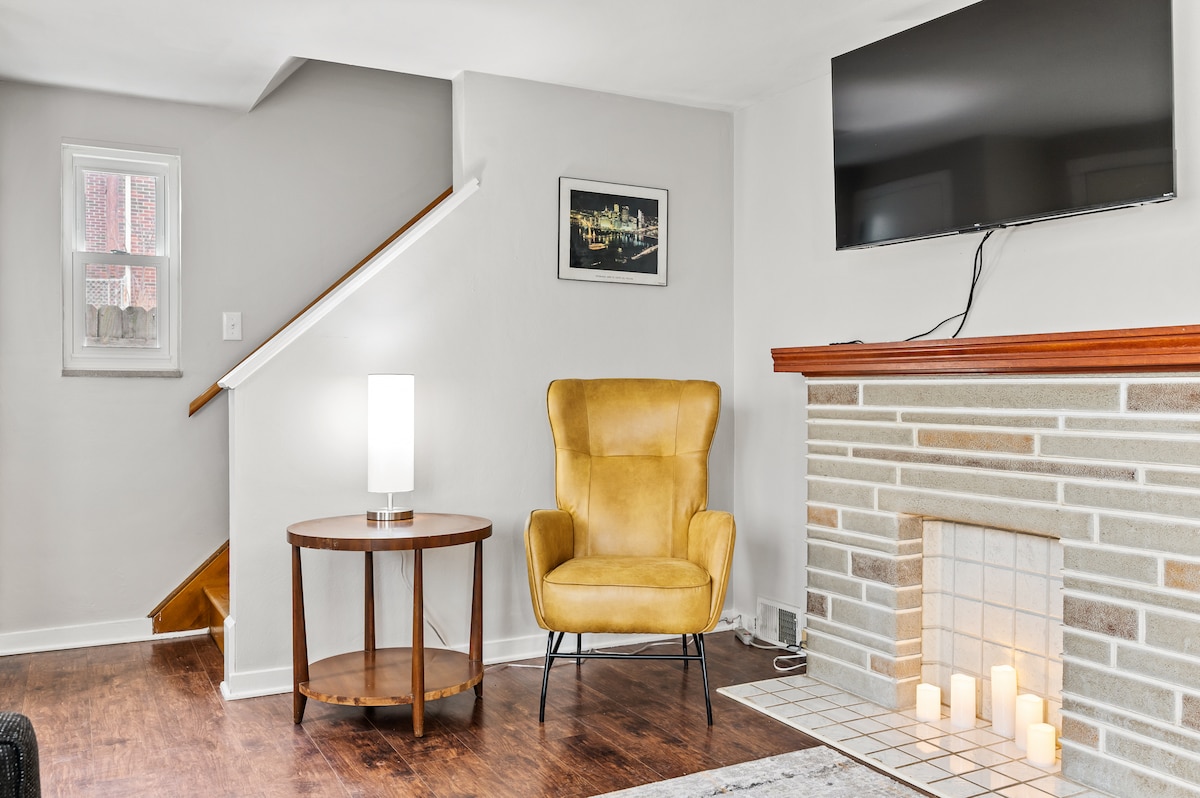 A comfortable living area is showcased with a modern yellow armchair positioned next to a round wooden table. A stone fireplace is present, along with a television mounted on the wall above. Light from a nearby lamp creates a cozy atmosphere.