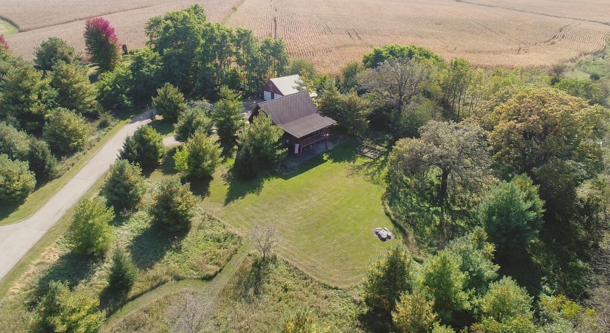 An aerial view showcases a secluded cabin surrounded by lush greenery, with open fields visible in the background. The spacious lawn in front of the cabin highlights the natural landscaping, providing a sense of tranquility. A winding road leads up to the property.