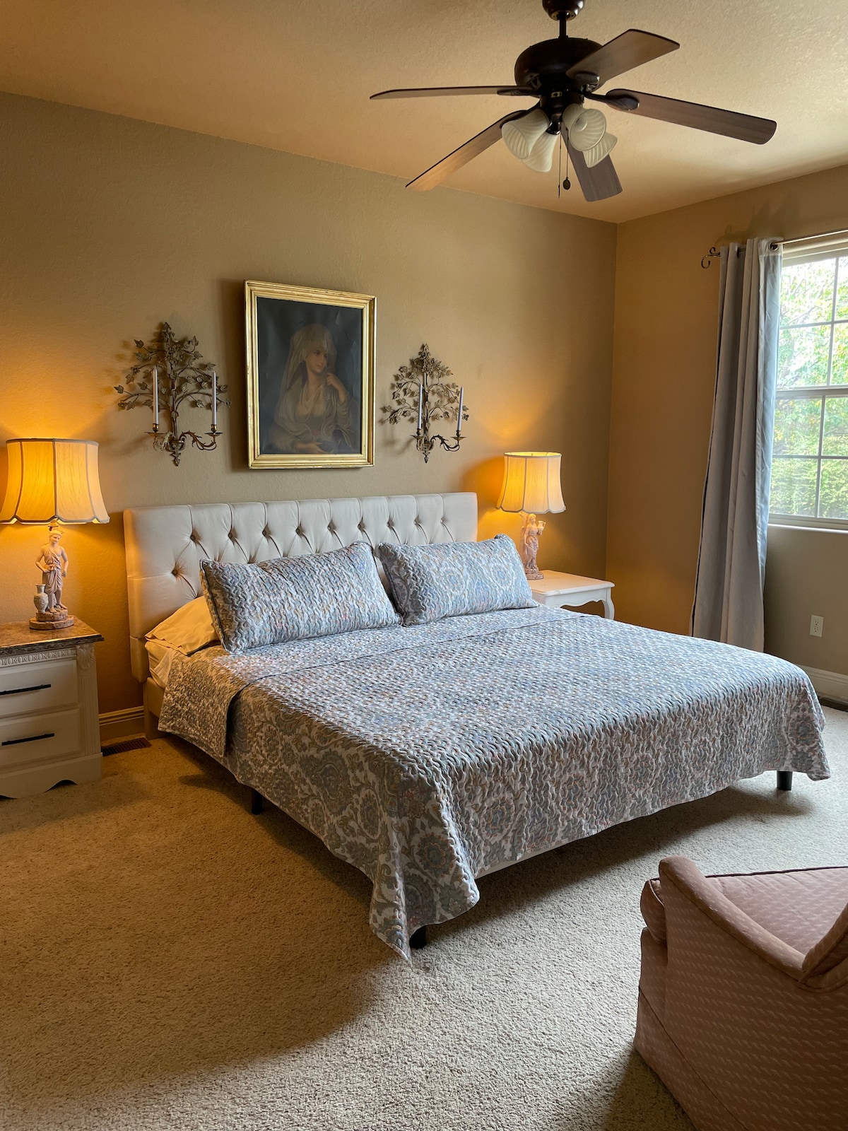A spacious bedroom features a plush bed with decorative bedding. Two bedside lamps provide soft illumination beside matching nightstands. A framed painting hangs above the bed, while natural light enters through a large window, complemented by light curtains. A cozy armchair is positioned in the corner.