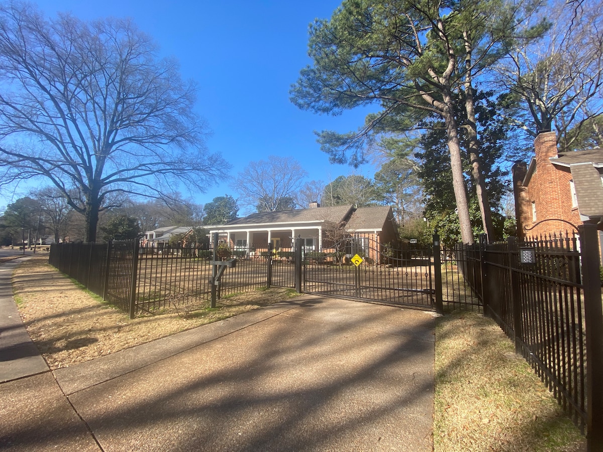 A gated entrance reveals a charming single-story home surrounded by trees. The property features a large porch with seating, situated behind a tall fence, creating a sense of privacy. A clear blue sky complements the inviting exterior.