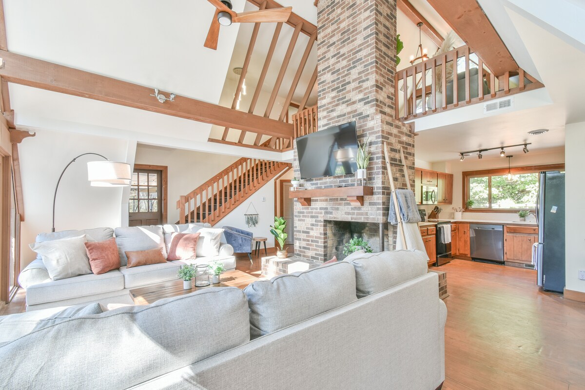 The interior living space features a comfortable sectional sofa adorned with decorative cushions, positioned near a stylish stone fireplace. A wooden staircase leads to an upper loft area, while large windows allow natural light to illuminate the open layout.