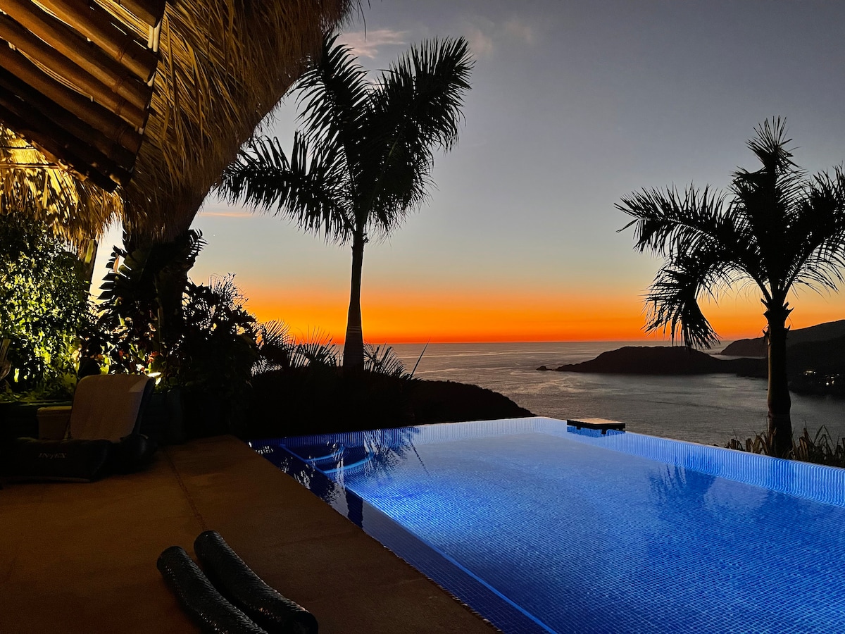 The infinity pool reflects the vibrant colors of the sunset, with a view overlooking the ocean and nearby hills. Palm trees frame the scene, adding a touch of nature. Soft lighting from the surrounding area creates a serene ambiance.