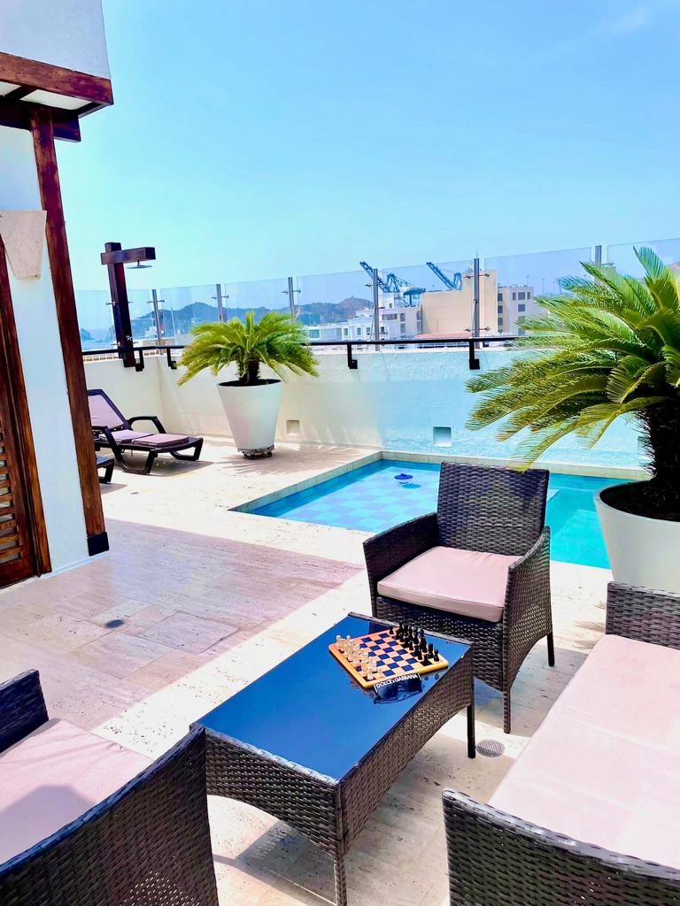 An outdoor space features a private pool surrounded by stylish lounge chairs and potted palm plants. A small table holds a chess set, offering a relaxed area for entertainment with views of the surrounding area and clear blue sky.