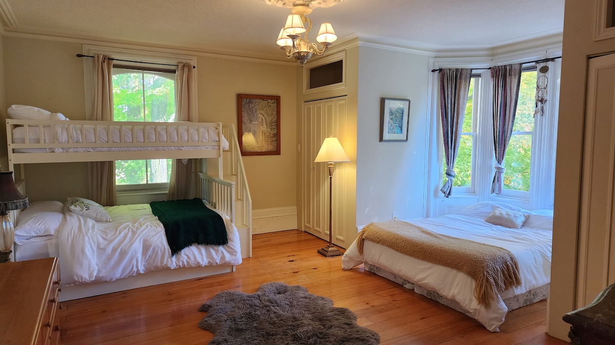 The Maple Room features a bunk bed with a single above a double bed, alongside a spacious queen bed. Natural light fills the room through large windows, highlighting the wooden flooring. A soft area rug is positioned on the floor, complemented by a bedside lamp and decorative artwork.