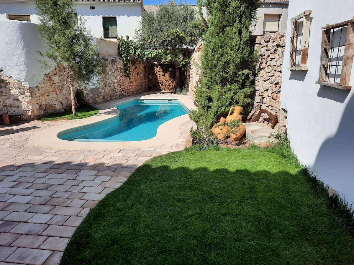 A private outdoor area features a rectangular swimming pool surrounded by stone walls and lush greenery. Neat patches of grass are visible alongside a tiled patio, which provides space for relaxation. Sunlight illuminates the area, creating a serene and inviting environment.