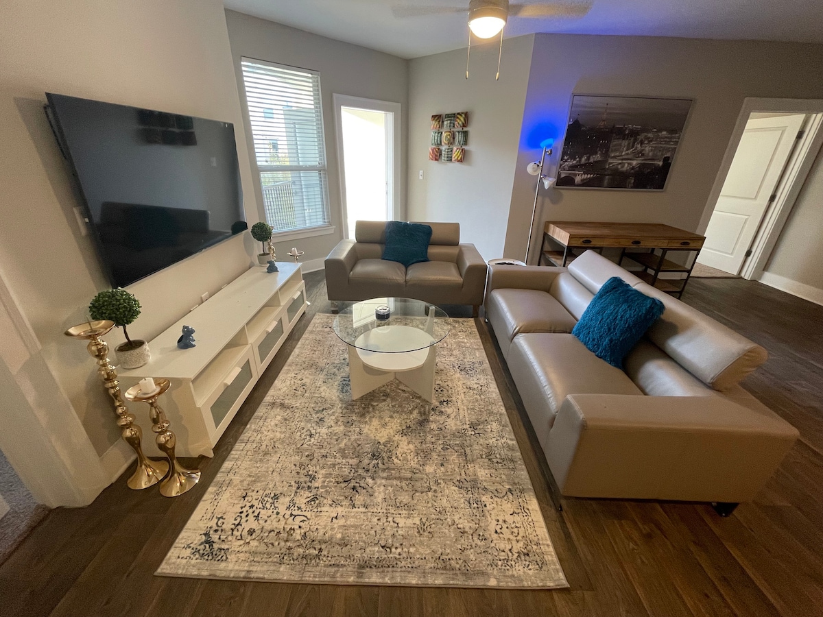A modern living area is highlighted by sleek, neutral-colored furniture. A glass coffee table sits atop a textured rug, while a large television is mounted on a white media console. Natural light enters through windows, and decorative accents include a small plant and wall art.