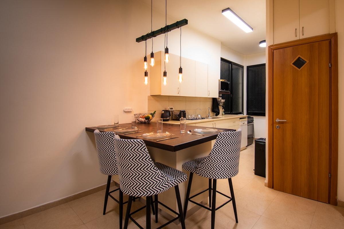 An open kitchen area is presented with a modern table featuring four patterned chairs. Pendant lights hang above the table, illuminating the space. A functional kitchen is visible in the background, equipped with essential appliances and a large window offering natural light.