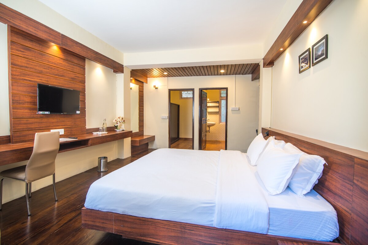 A spacious bedroom features a wooden accent wall and a comfortable bed with white linens. A desk with a chair is positioned against one wall, complemented by soft lighting fixtures. A television is mounted nearby, and light flows in from an adjacent doorway.