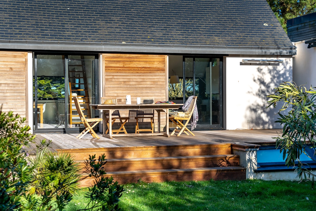 A spacious wooden terrace features a dining table surrounded by folding chairs. Glass doors offer a view into the interior space, while the lush green lawn and plants outline the area, providing a natural setting for relaxation.