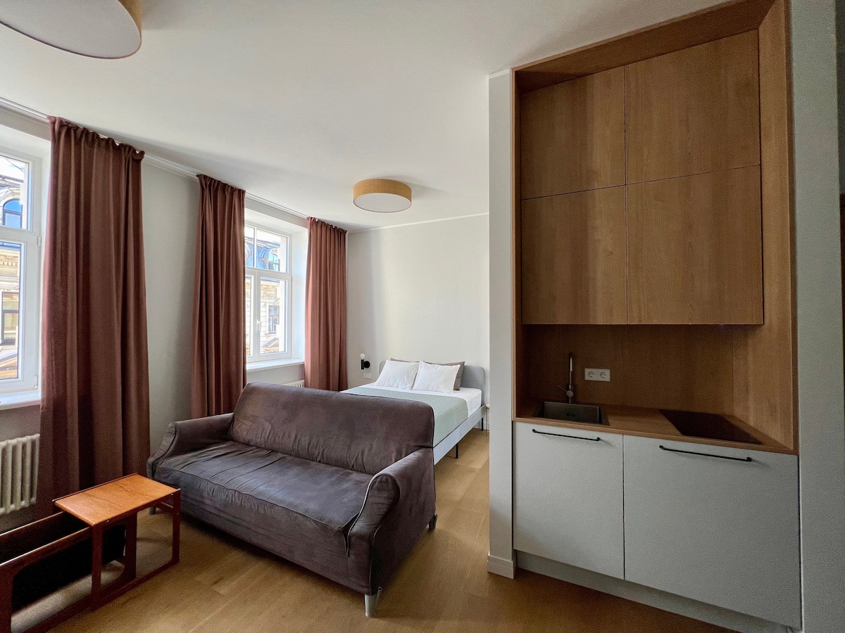 A bright studio space is featured, showcasing a spacious bed with white linens. A comfortable gray sofa is positioned nearby, accompanied by a small wooden table. Natural light enters through two windows, framed by soft brown curtains, while a minimalist kitchenette is visible in the corner.