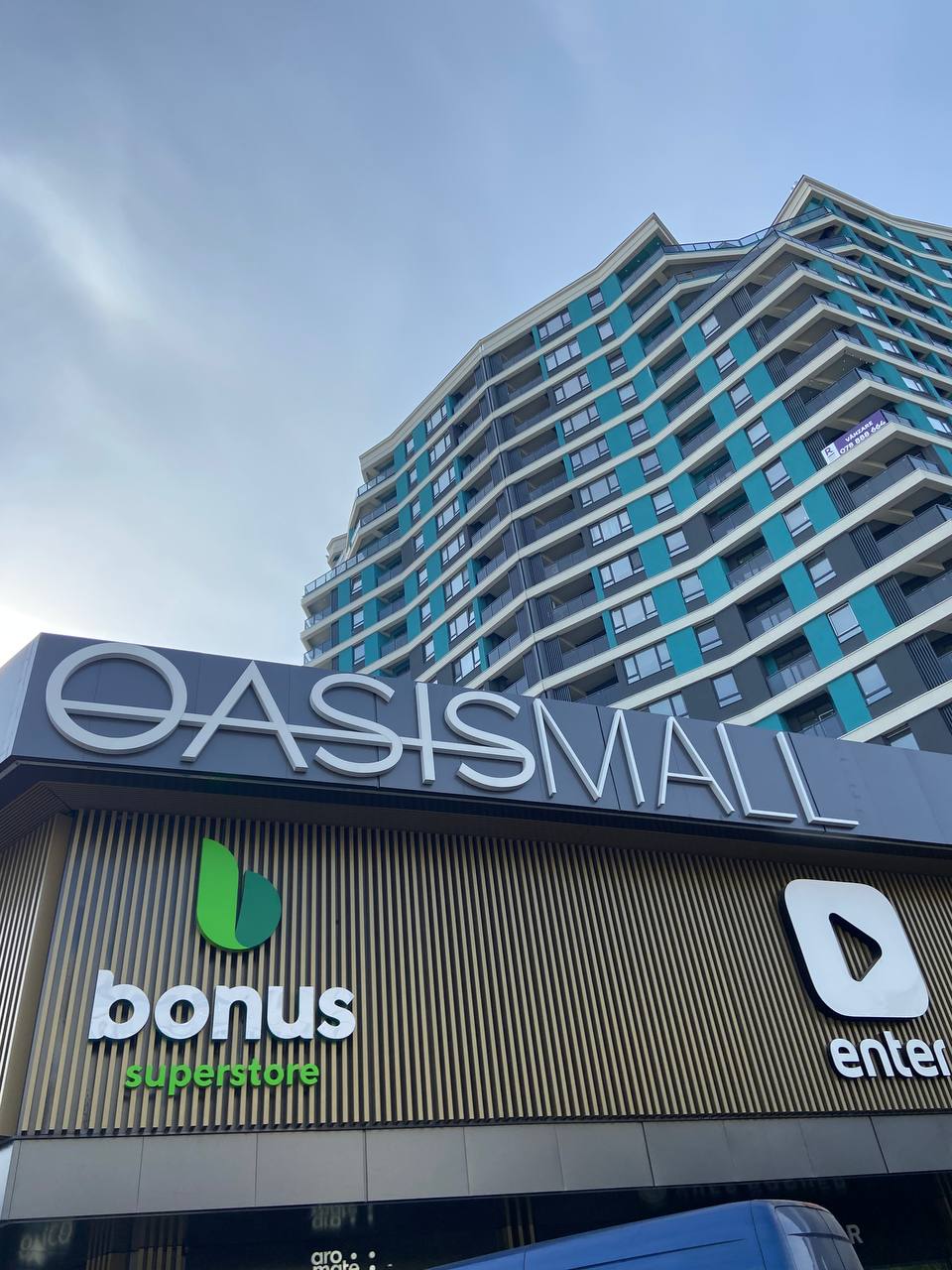 The exterior of a modern building features the name 'OASISMALL' prominently displayed. Below, a storefront is labeled 'bonus superstore,' while a recognizable logo is visible next to it. The design reflects contemporary architecture with a blend of blue and white elements.