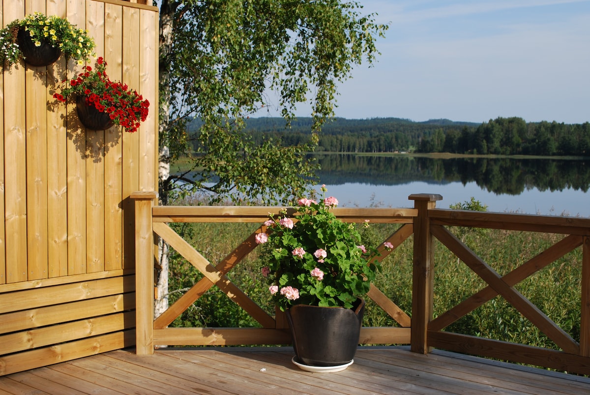 A wooden deck is framed by a natural wooden railing and features flower pots with vibrant blooms. In the background, serene water reflects the surrounding greenery, creating a calm atmosphere. The unobstructed view highlights the diverse landscape beyond the deck.
