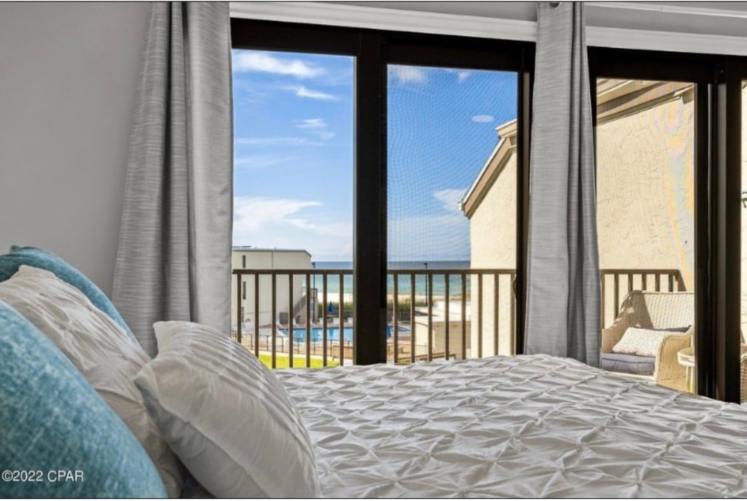 A spacious bedroom features a comfortable bed with soft bedding. Large glass doors open to a private balcony, allowing for views of the ocean and the pool area. Natural light fills the room, accentuating the elegant curtains.
