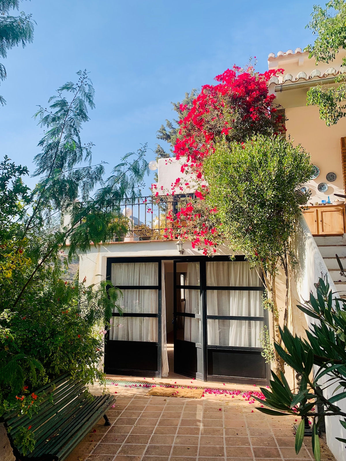 Two sets of black double doors are framed by vibrant bougainvillea and lush greenery. A tiled pathway leads up to the entrance, and a small balcony is visible above. Sunlight illuminates the area, creating a cheerful and welcoming atmosphere.
