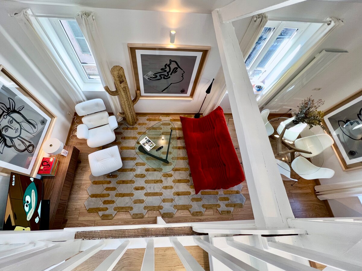 An overhead view of the living area shows a spacious layout with contemporary furnishings. A red couch and white armchairs are arranged on a patterned rug, complemented by large artworks on the walls. Natural light floods the space through multiple windows.