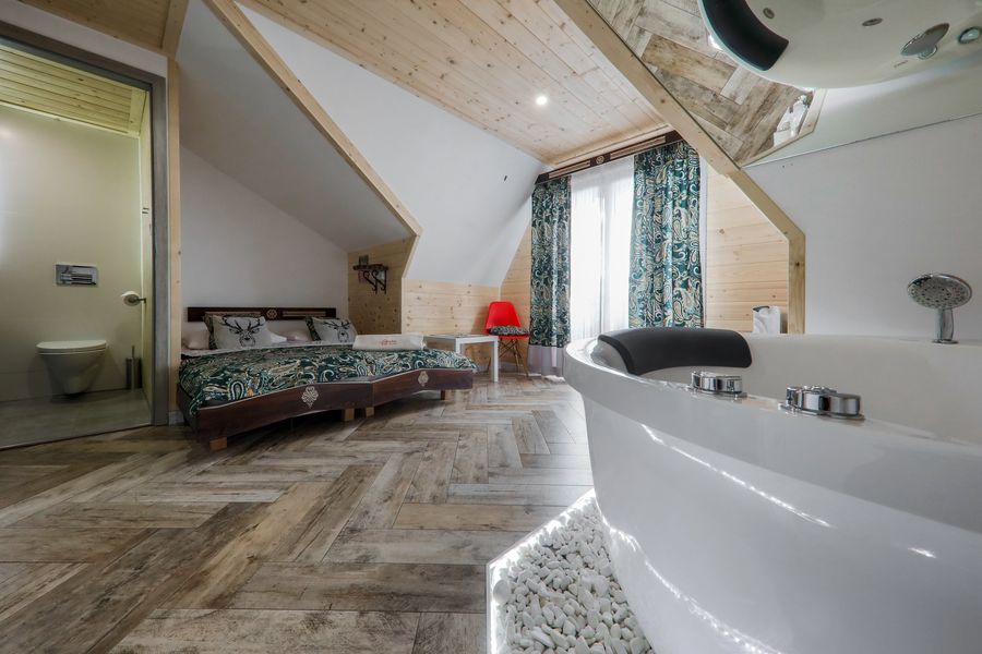 A spacious room features wooden paneling and a sloped ceiling. A comfortable bed is complemented by a colorful bedspread, while a modern bathtub is positioned nearby on a bed of decorative stones. A window adorned with patterned curtains allows natural light to fill the space.