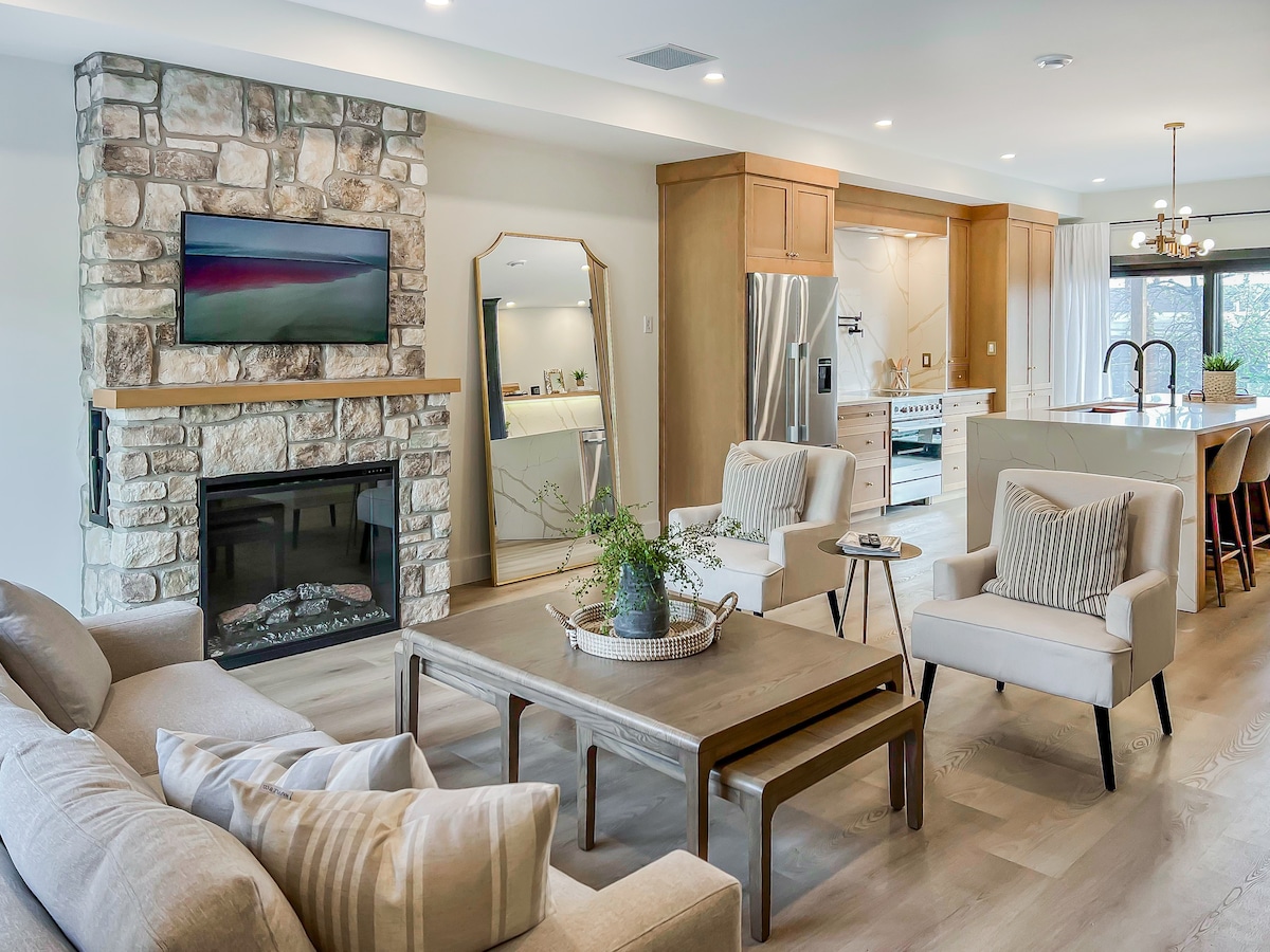 A spacious living area features a stone fireplace with a mounted TV above it. Comfortable seating is provided by a light-colored sofa and two armchairs. A wooden coffee table occupies the center, surrounded by neutral-toned décor. Bright light flows through the nearby windows, enhancing the warm atmosphere.