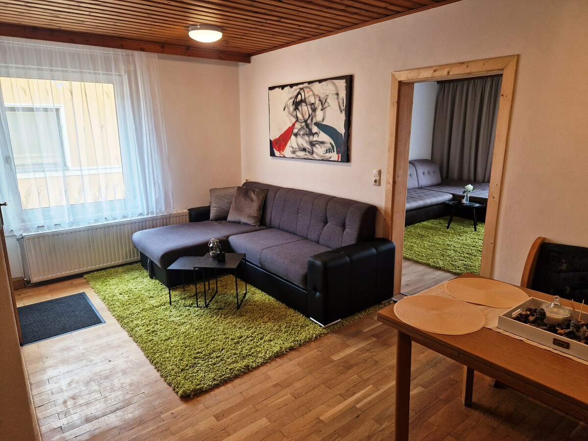 A cozy living area features a comfortable dark-colored sofa beside a small black coffee table, set on a vibrant green rug. Light filters in through a large window dressed with sheer curtains. An adjacent dining area includes a wooden table with decorative plates.