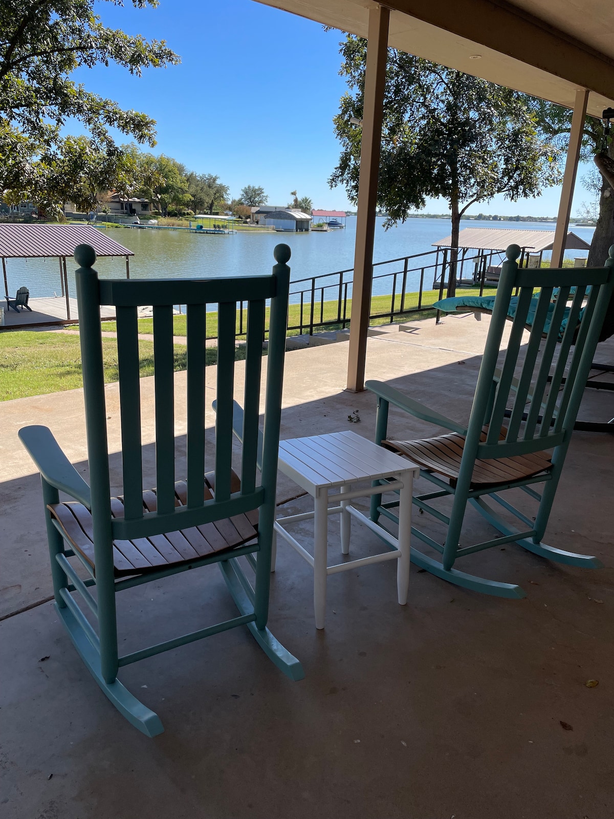 Two rocking chairs are positioned on a spacious patio overlooking Lake Nasworthy. A small white table is placed between them, offering a spot for drinks. The view includes calm waters and greenery, creating a serene outdoor space for relaxation.
