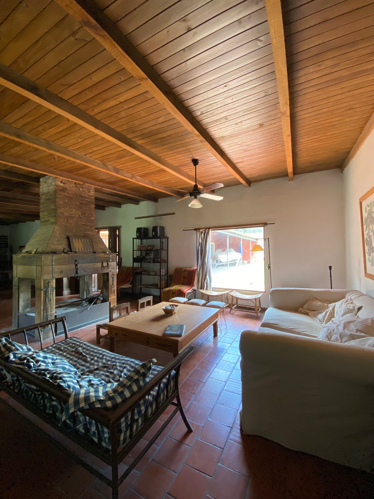 A spacious living area features a wooden ceiling with exposed beams. A comfortable white sofa and a patterned seating area are arranged around a wooden coffee table. A lamp stands nearby, and large doors allow natural light to fill the room.