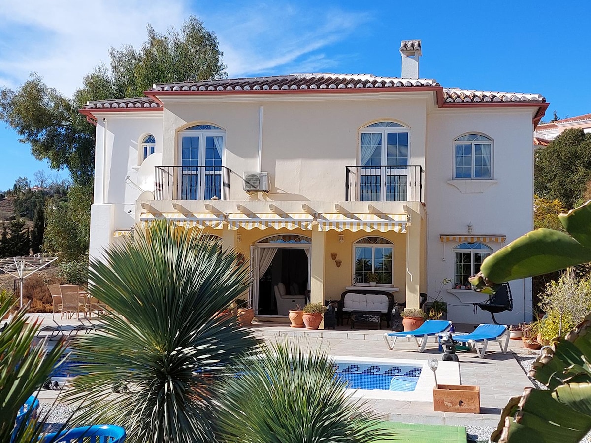 The villa features a light-colored exterior with expansive windows and a balcony. Surrounding greenery adds a natural touch. An inviting pool is visible in the front, accompanied by lounge chairs and potted plants, creating a serene outdoor space.