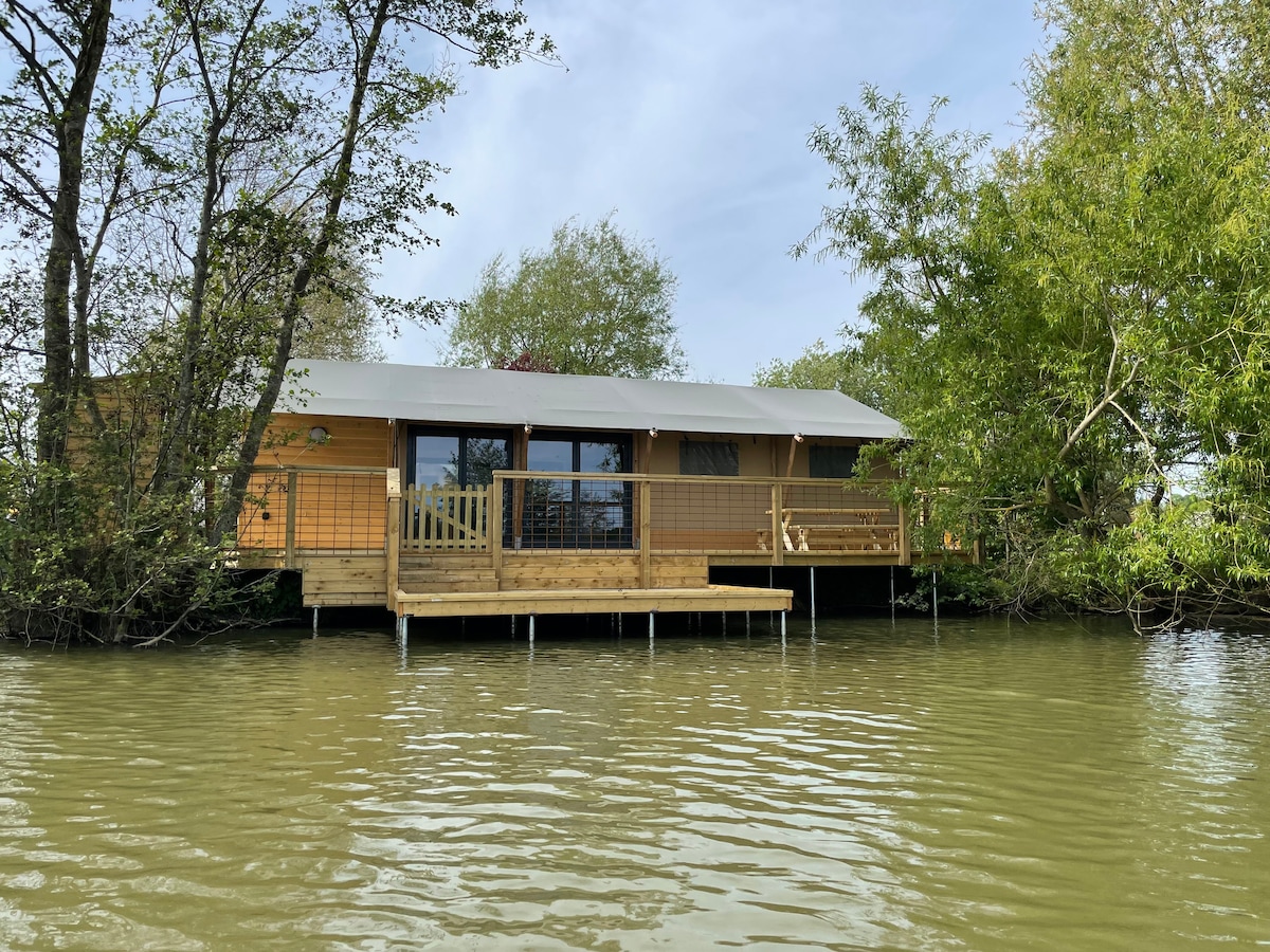 A wooden Safari Tent is positioned on stilts above the calm waters of Sumners Lake, surrounded by lush greenery. Large windows provide a view of the natural landscape, while a deck extends outwards for outdoor relaxation.