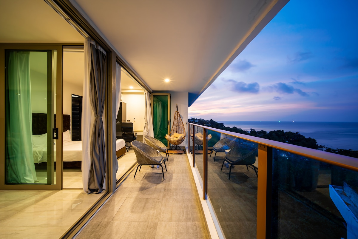 A spacious balcony overlooks a tranquil ocean view, where the sky transitions to evening hues. Two comfortable chairs are positioned for relaxation, and glass doors provide access to the interior living space. Soft lighting enhances the inviting atmosphere as dusk approaches.